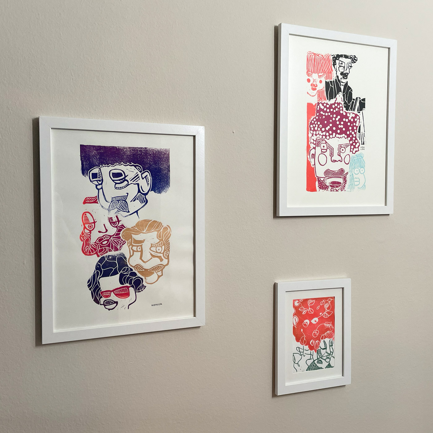 Three colorful linoprint artworks with abstract faces in white frames on beige wall